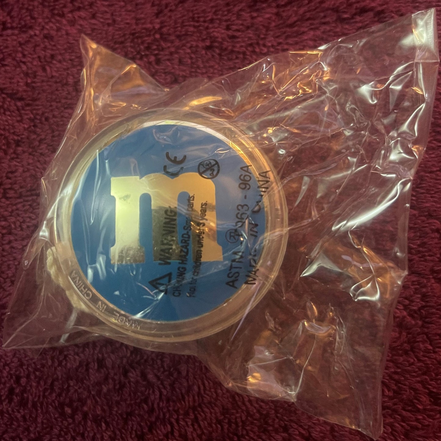 Sealed clear plastic package containing blue and yellow M&M's yo-yo with visible branding, displayed against maroon fabric ba