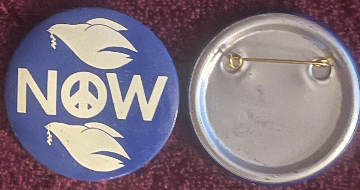Front and back of blue pinback button with cream dove and "NOW" text with peace symbol, shown against burgundy fabric
