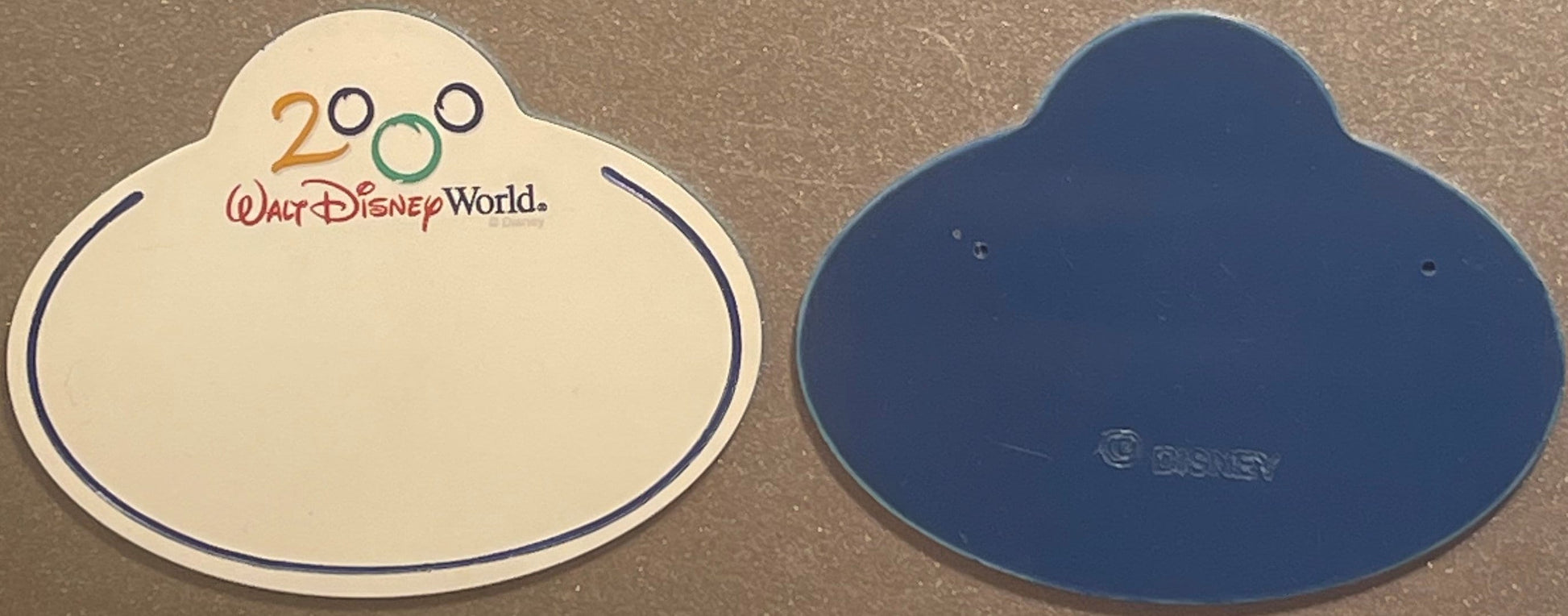 Front and back of a cream and blue Mickey-shaped Disney World 2000 cast member badge with "Walt Disney World" text and Disney