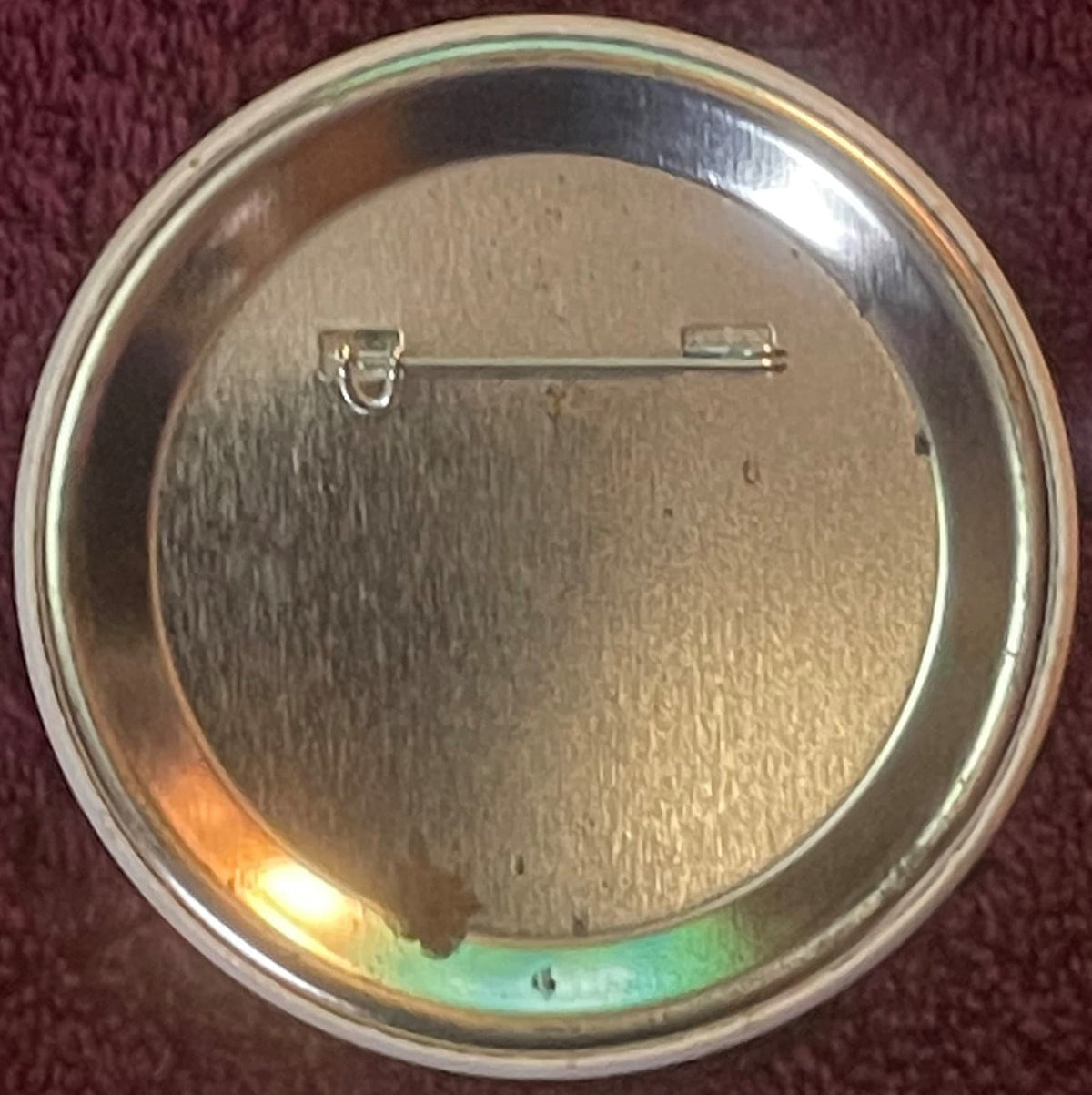 Back of metal pin badge showing safety clasp against maroon fabric background, with green and gold coloring visible on rim