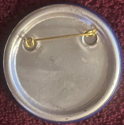 Back of vintage pinback button with gold-toned pin and clasp on cream-colored circular backing, displayed on burgundy fabric