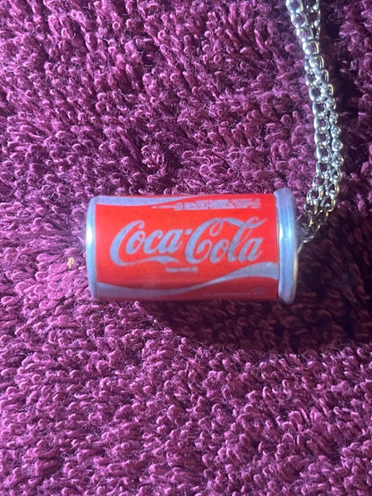 Sealed miniature Coca-Cola can with red label and white ends on purple textured fabric, with silver chain visible at right ed