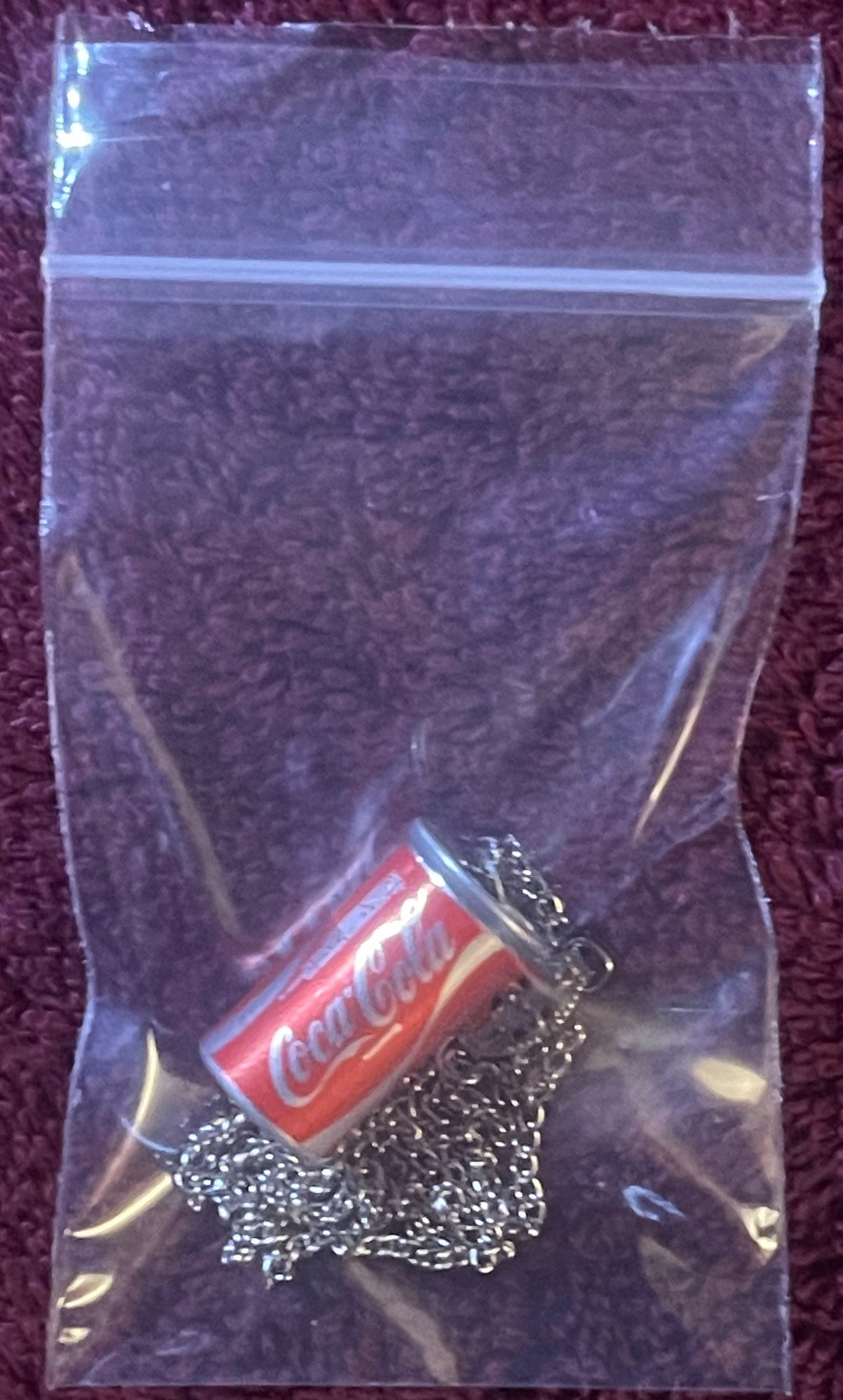 Small red Coca-Cola can pendant on silver chain in clear zip-lock bag against purple fabric background