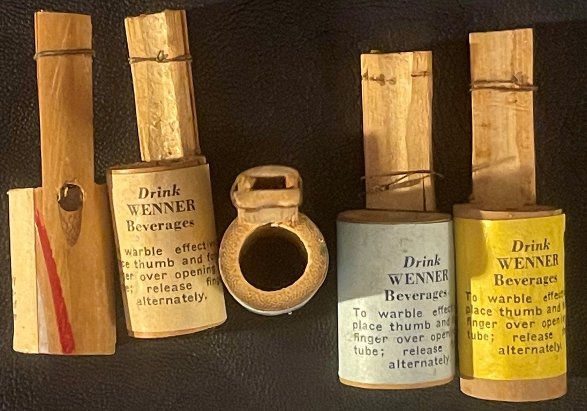 Five vintage wooden Wenner Beverages Warbler novelty whistles with instructional labels, arranged on dark fabric for display.
