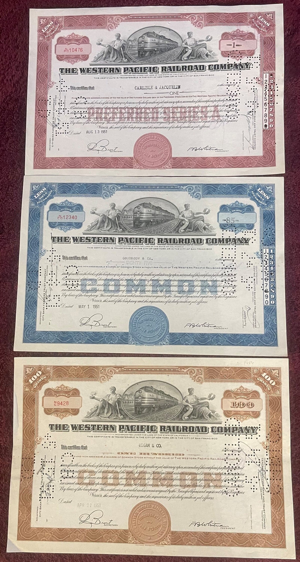 Three Western Pacific Railroad Company stock certificates in pink, blue, and brown, dated 1951, displayed on burgundy fabric 