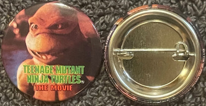 Front and back of a circular pin featuring Michelangelo from Teenage Mutant Ninja Turtles movie, with green text logo on oran