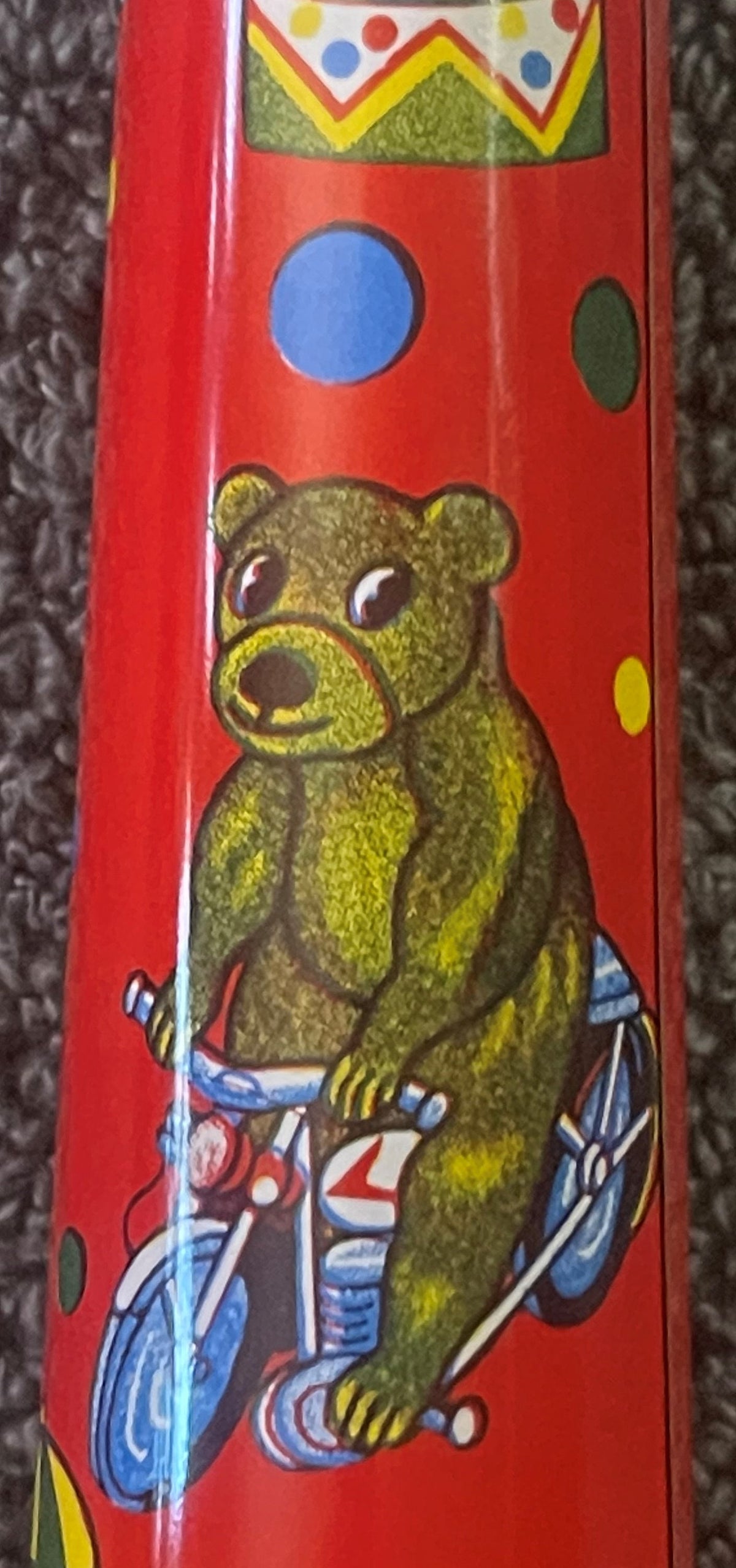 Vintage red tin circus horn noisemaker with yellow bear on tricycle, colorful dots and crown design at top, shown against gra