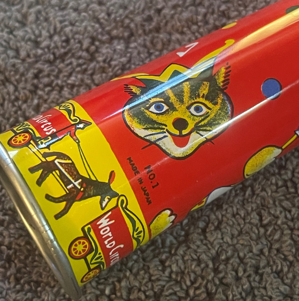 Vintage red tin horn noisemaker with yellow and black lithographed circus animal designs, shown at an angle on gray carpet