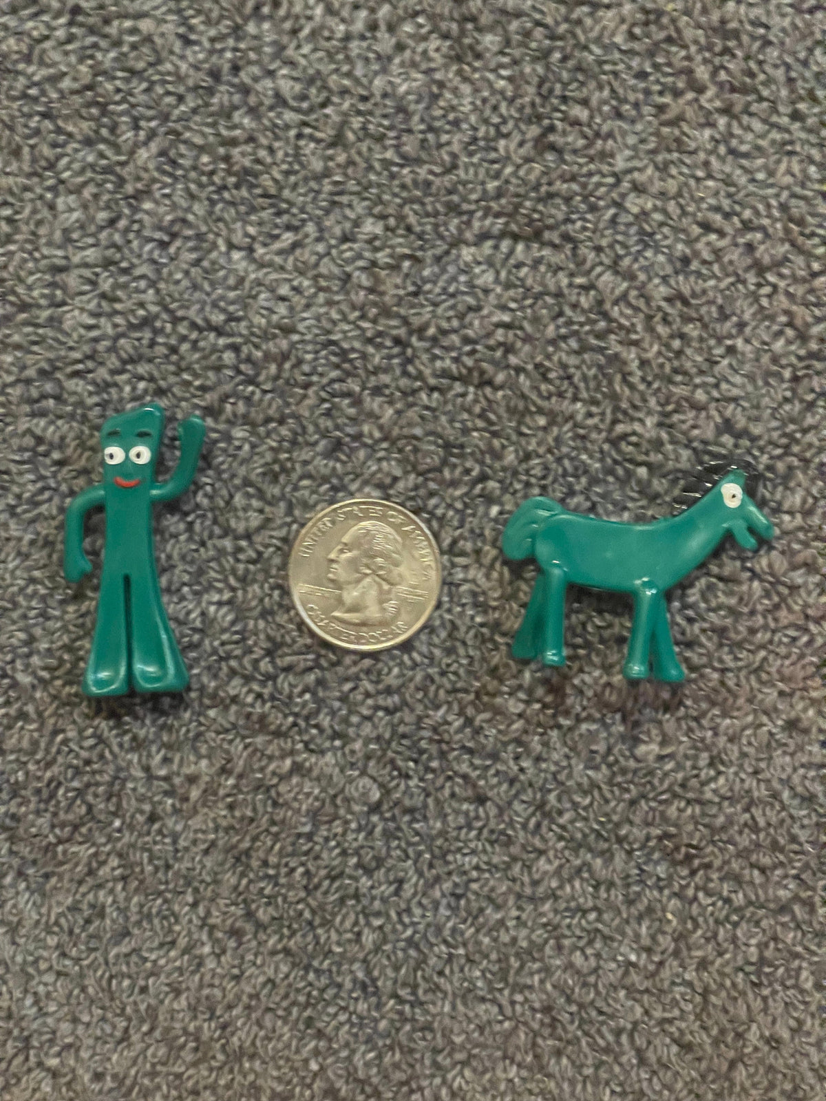 A teal Gumby and a teal Pokey horse laid beside a US quarter for scale on gray textured fabric.
