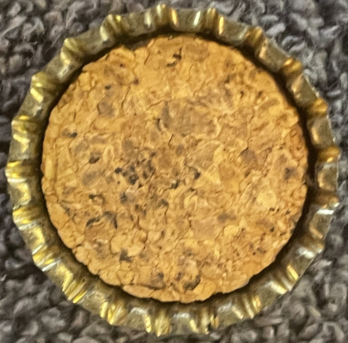 Antique Ritz Orange Pineapple Soda cork bottle cap with gold-ridged edge, tan cork center with visible dark specks, photographed flat on gray textured fabric background.