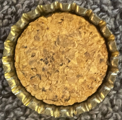 Antique Ritz Orange Pineapple Soda cork bottle cap with gold-ridged edge, tan cork center with visible dark specks, photographed flat on gray textured fabric background.