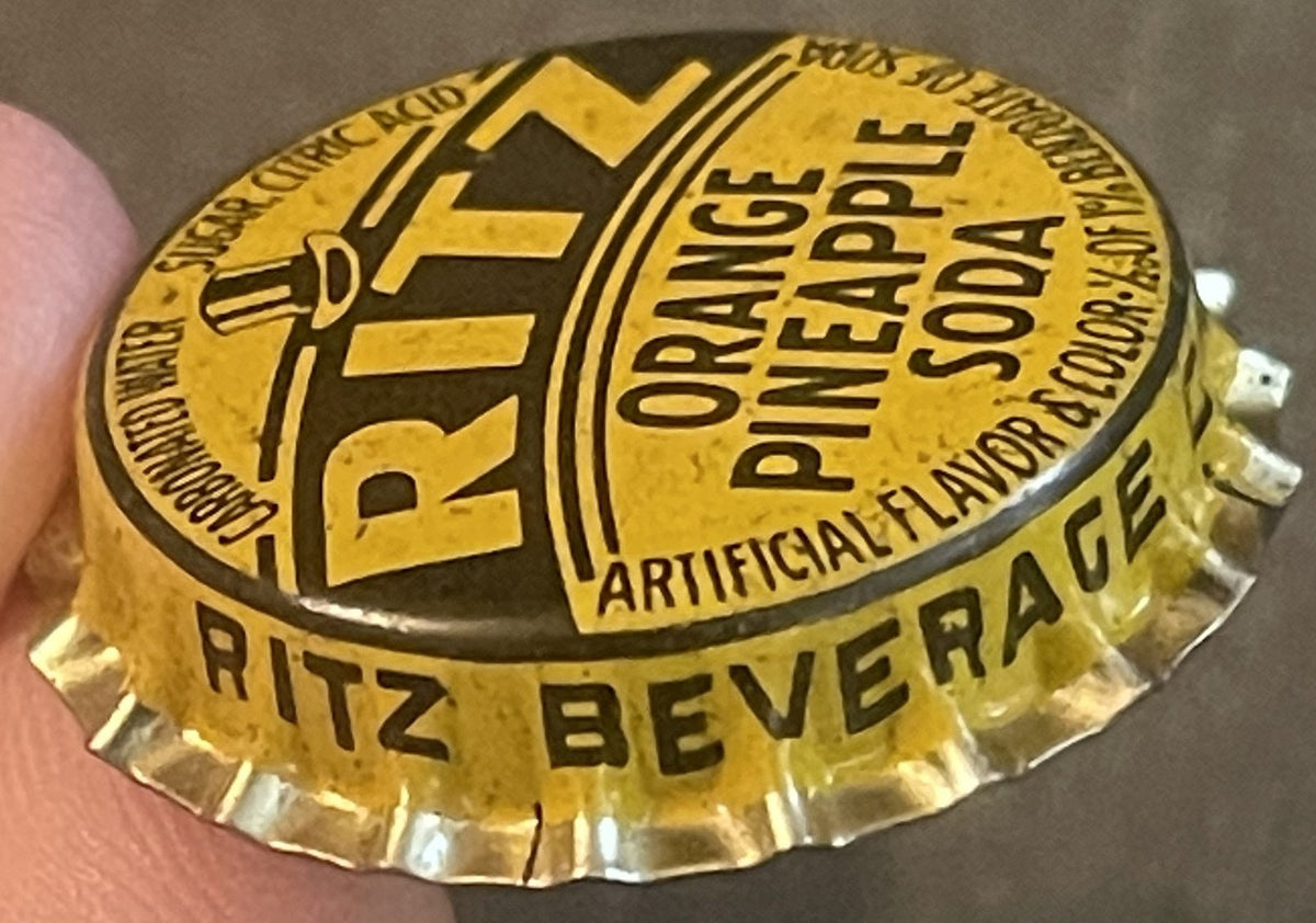 Antique Ritz Orange Pineapple Soda bottle cap, gold with brown embossing, held in hand for scale. Text reads "Ritz Beverage," "Orange Pineapple Soda," and "Artificial Flavor," St. Louis MO.