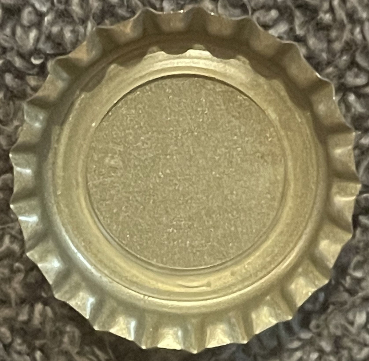 Vintage metal bottle cap with scalloped edges and embossed circular rim, shown flat against textured fabric background