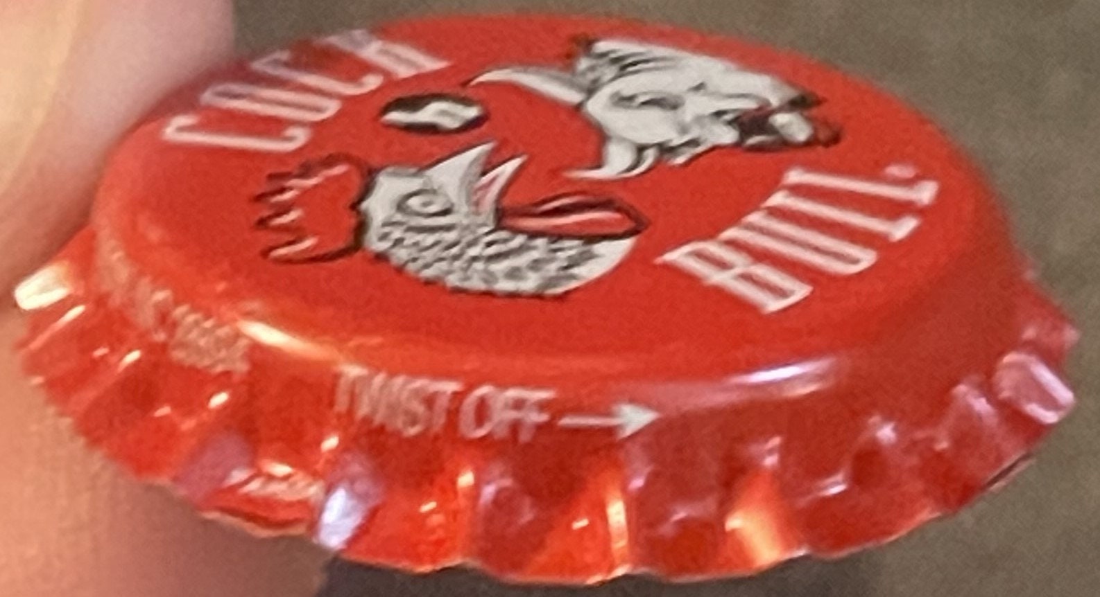 Red vintage bottle cap with embossed rooster and bull graphics, held in hand showing ridged edges and metallic lettering
