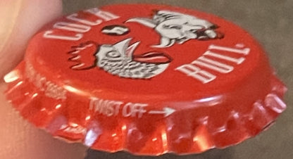 Red vintage bottle cap with embossed rooster and bull graphics, held in hand showing ridged edges and metallic lettering