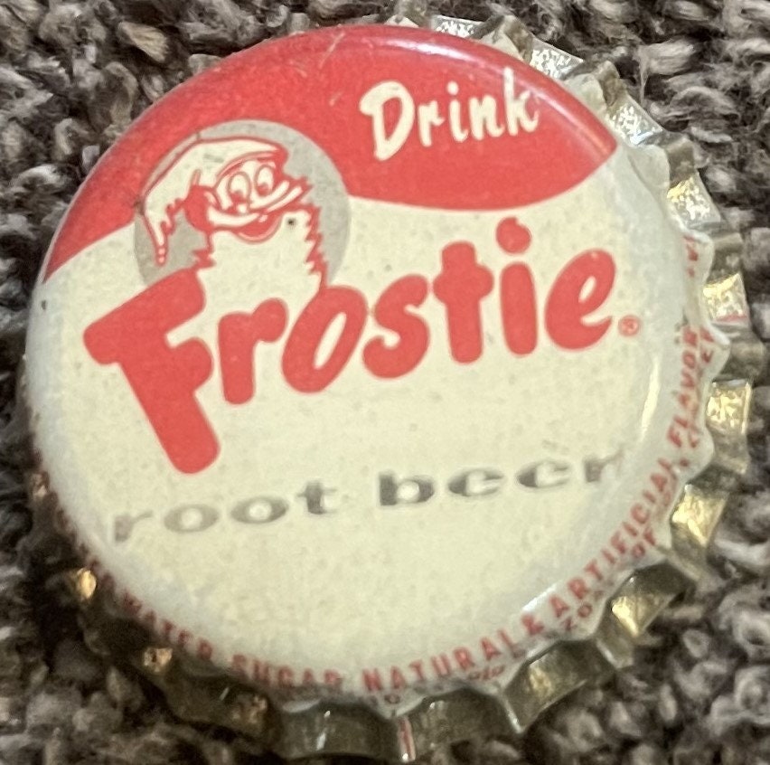 Vintage Frostie Root Beer cork-lined bottle cap featuring red and white design with elf mascot logo and "Drink Frostie Root Beer" text, shown flat against textured fabric background.