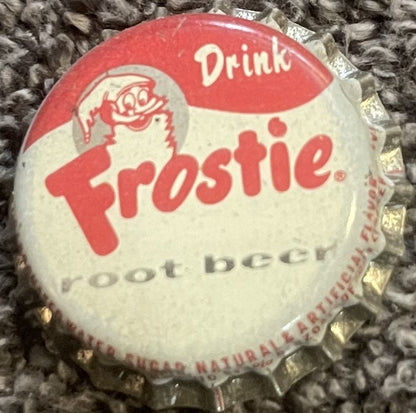 Vintage Frostie Root Beer cork-lined bottle cap featuring red and white design with elf mascot logo and "Drink Frostie Root Beer" text, shown flat against textured fabric background.
