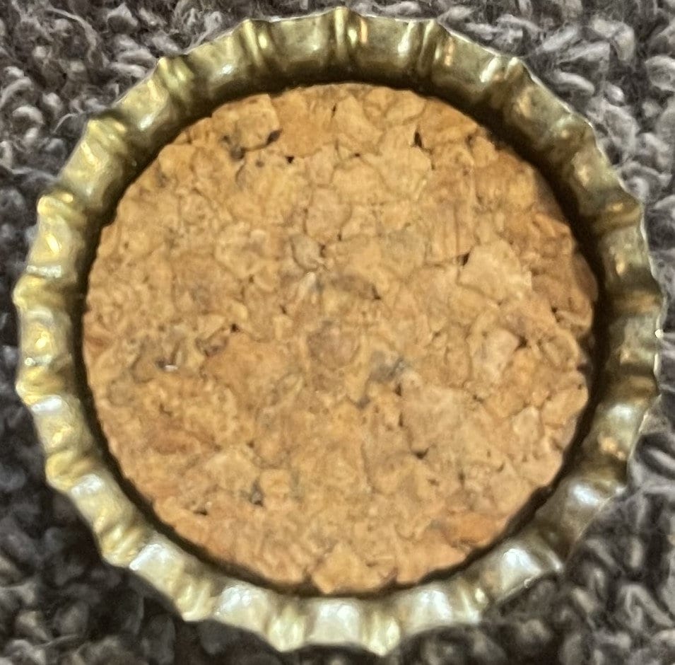 Top-down view of the inside of a crown cap with a tan granulated cork liner and a fluted gold-toned metal rim on gray fabric.
