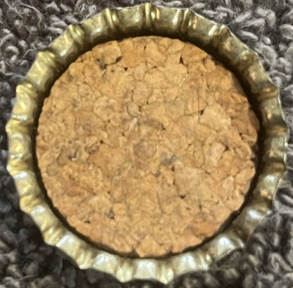 Top-down view of the inside of a crown cap with a tan granulated cork liner and a fluted gold-toned metal rim on gray fabric.