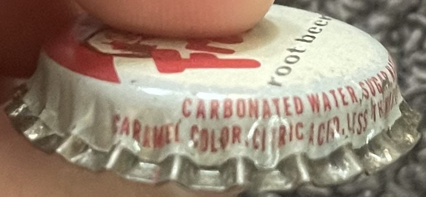 Side view of a Frostie root beer cap held by a finger, with red text on the skirt reading 'Carbonated Water, Sugar, Caramel Color, Citric Acid'.