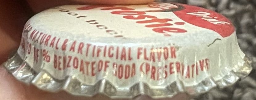 Side view of a Frostie root beer cap held by a hand, with red skirt text 'Natural & Artificial Flavor' and 'Benzoate of Soda (Preservative)'.