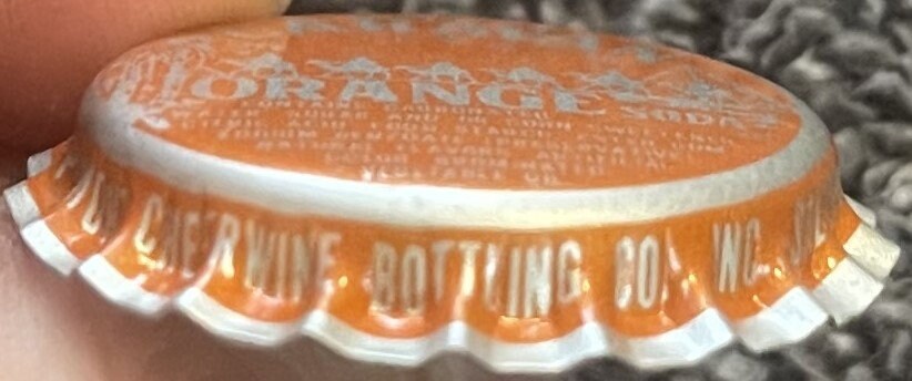 Vintage orange and white bottle cap held in hand showing embossed text and ridged edges, close-up detail view