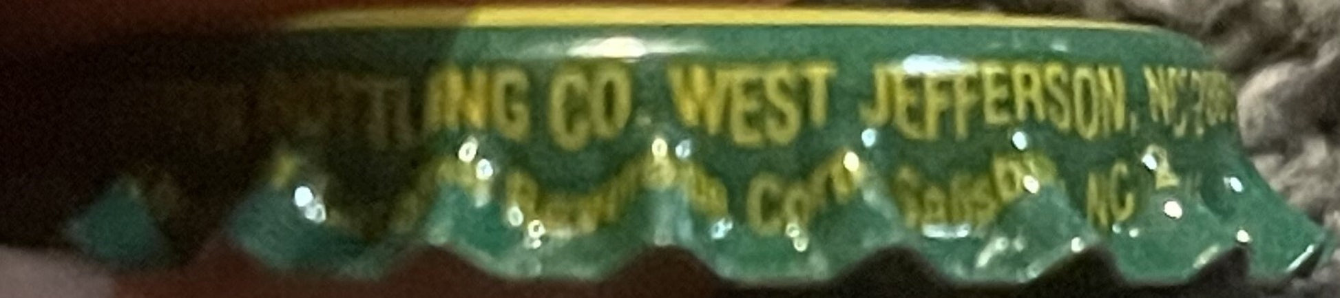 Close-up of a green and gold Cheerwine twist-off bottle cap showing embossed text including "West Jefferson" and bottler info