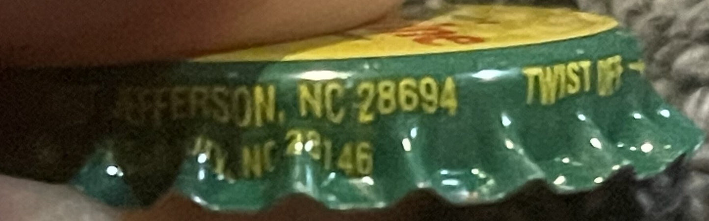 Close-up of green twist-off bottle cap with yellow text reading "JEFFERSON, NC 28694" and partial visible text, held by a han