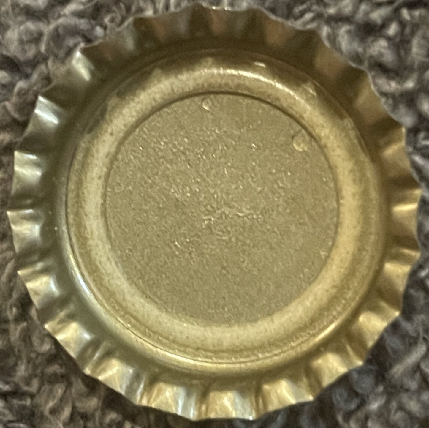Vintage gold twist-off bottle cap with ridged edges, shown flat against gray textured surface