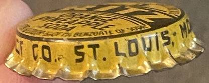 Antique Ritz Orange Pineapple soda bottle cap from St. Louis, MO, shown close-up held by a thumb, displaying embossed text and scalloped edges on gold-colored metal.
