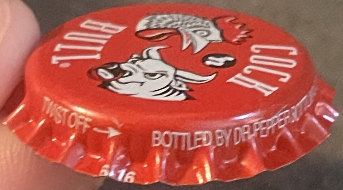 Red bottle cap with rooster and bull head graphics, held in a person's hand, text reads "Cock 'n Bull" and "Bottled By