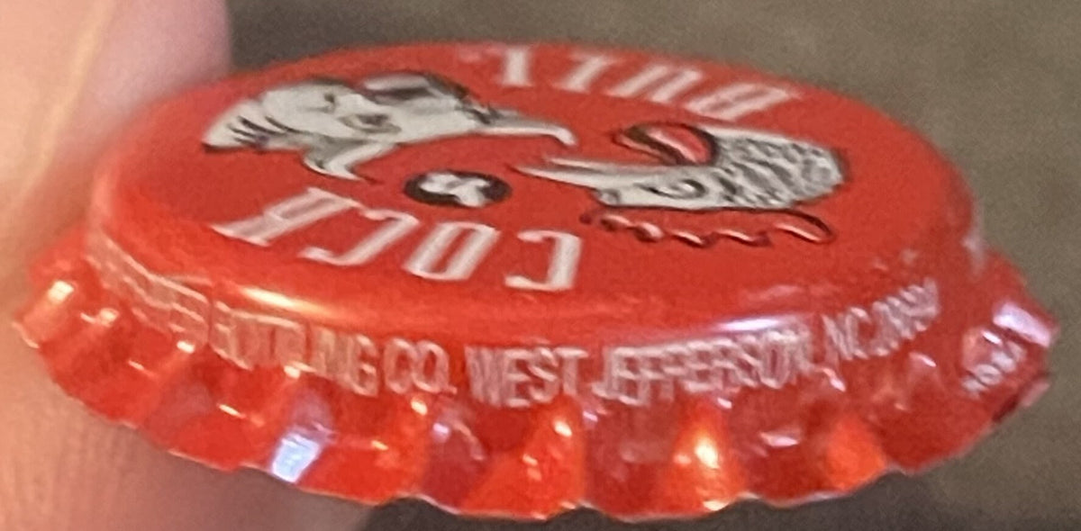 Red vintage bottle cap with rooster and bull graphics, held in hand showing embossed text and metallic shine detail