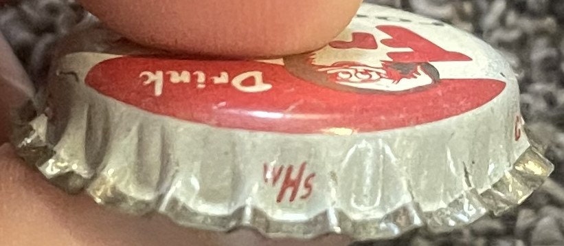 Side view of a Frostie root beer crown cap held between fingers: the red Drink band and elf upside down, and the small WHs cap-maker mark on the skirt.