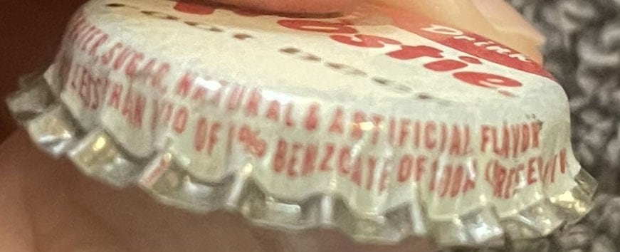 Side view of a Frostie root beer cap held by a hand, with red skirt text 'Natural & Artificial Flavor, Less than 1/10 of 1% Benzoate of Soda'.