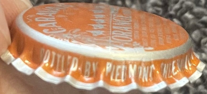 Vintage orange soda bottle cap held in hand showing embossed sun design and text, white ridged skirt visible