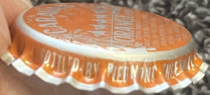 Vintage orange soda bottle cap held in hand showing embossed sun design and text, white ridged skirt visible