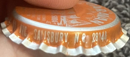 Close-up of orange bottle cap with white ridged edges, held by a hand, showing embossed text including "SALISBURY N.C. 28144