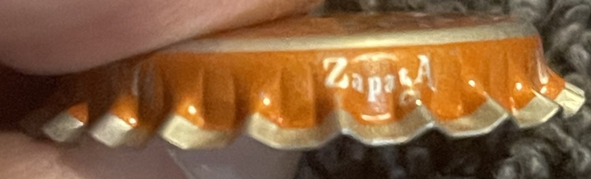 Close-up of orange soda bottle cap with white ridged edges and "Zappa" text visible, held by a finger for scale