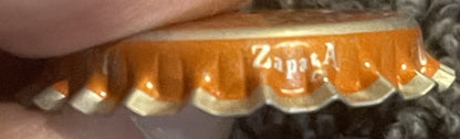 Close-up of orange soda bottle cap with white ridged edges and "Zappa" text visible, held by a finger for scale