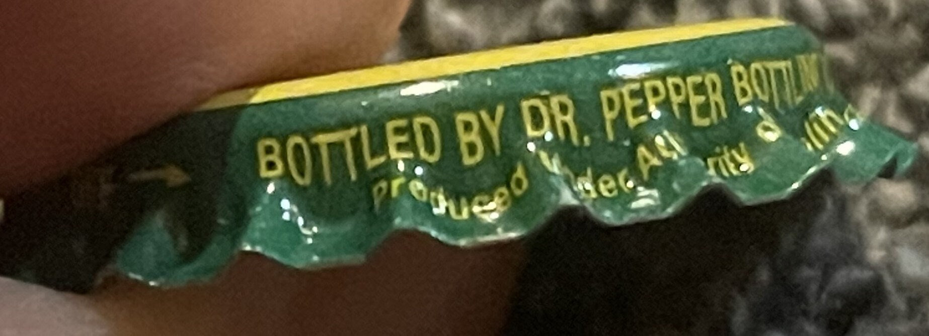 Close-up of green and yellow twist-off bottle cap with "BOTTLED BY DR. PEPPER BOTTLING" text, held by a hand for scale