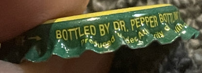 Close-up of green and yellow twist-off bottle cap with "BOTTLED BY DR. PEPPER BOTTLING" text, held by a hand for scale