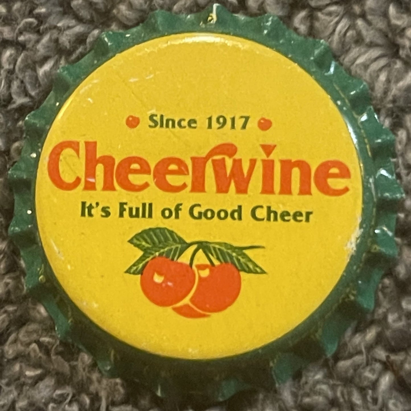 Vintage Cheerwine bottle cap on textured fabric background, yellow center with red cherries and green ridged edges, "Since 19