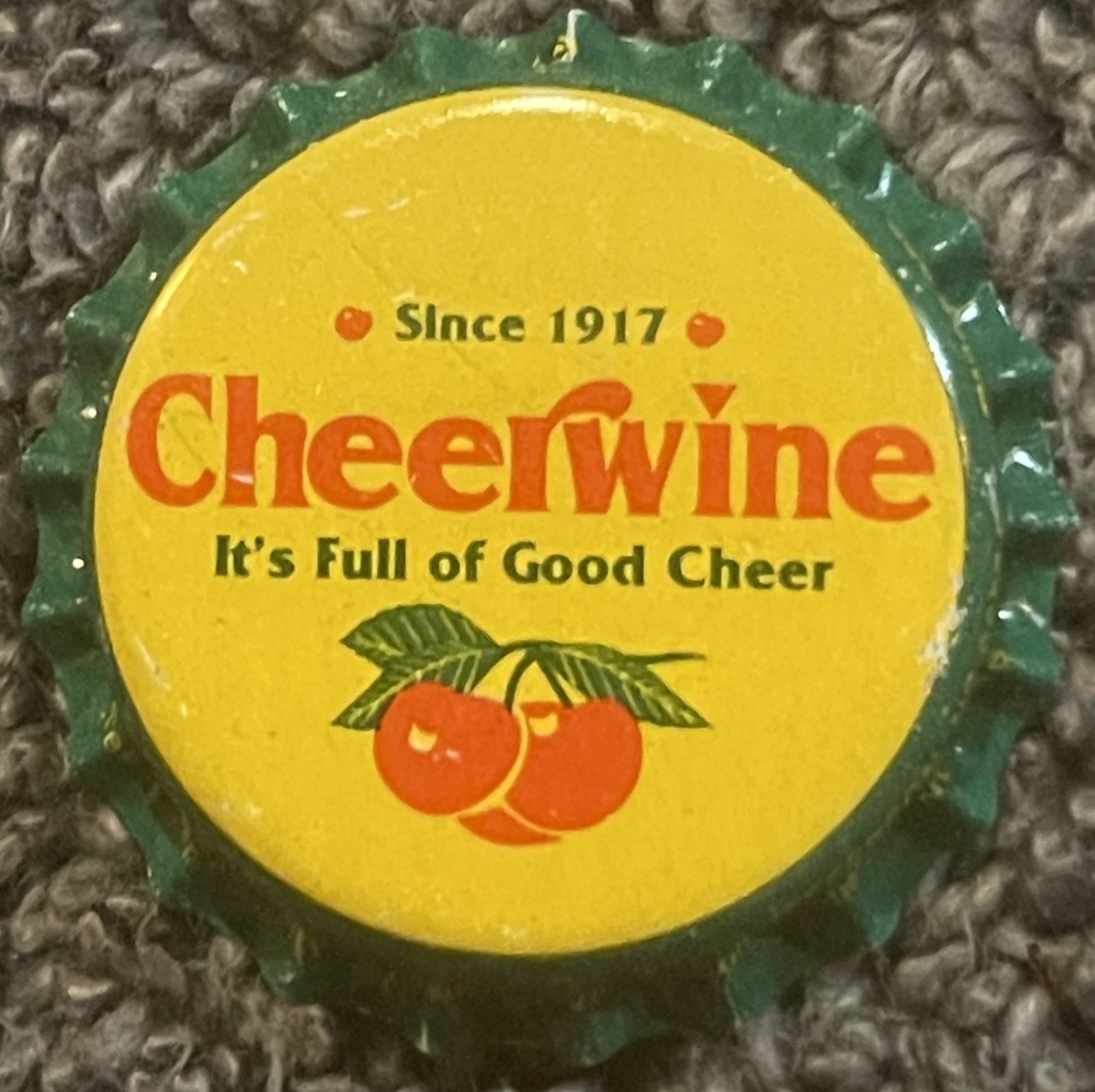 Vintage Cheerwine bottle cap on textured fabric background, yellow center with red cherries and green ridged edges, "Since 19