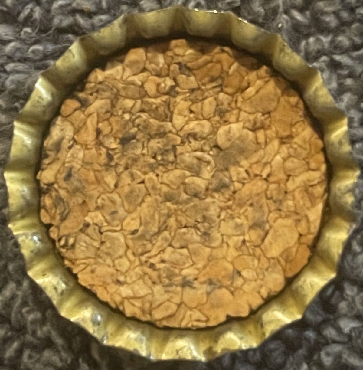 Antique Hires Root Beer cork bottle cap viewed from above, showing tan cork center with embossed pattern and yellow-gold crimped metal rim, on textured gray fabric background.