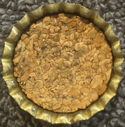 Antique Hires Root Beer cork bottle cap viewed from above, showing tan cork center with embossed pattern and yellow-gold crimped metal rim, on textured gray fabric background.