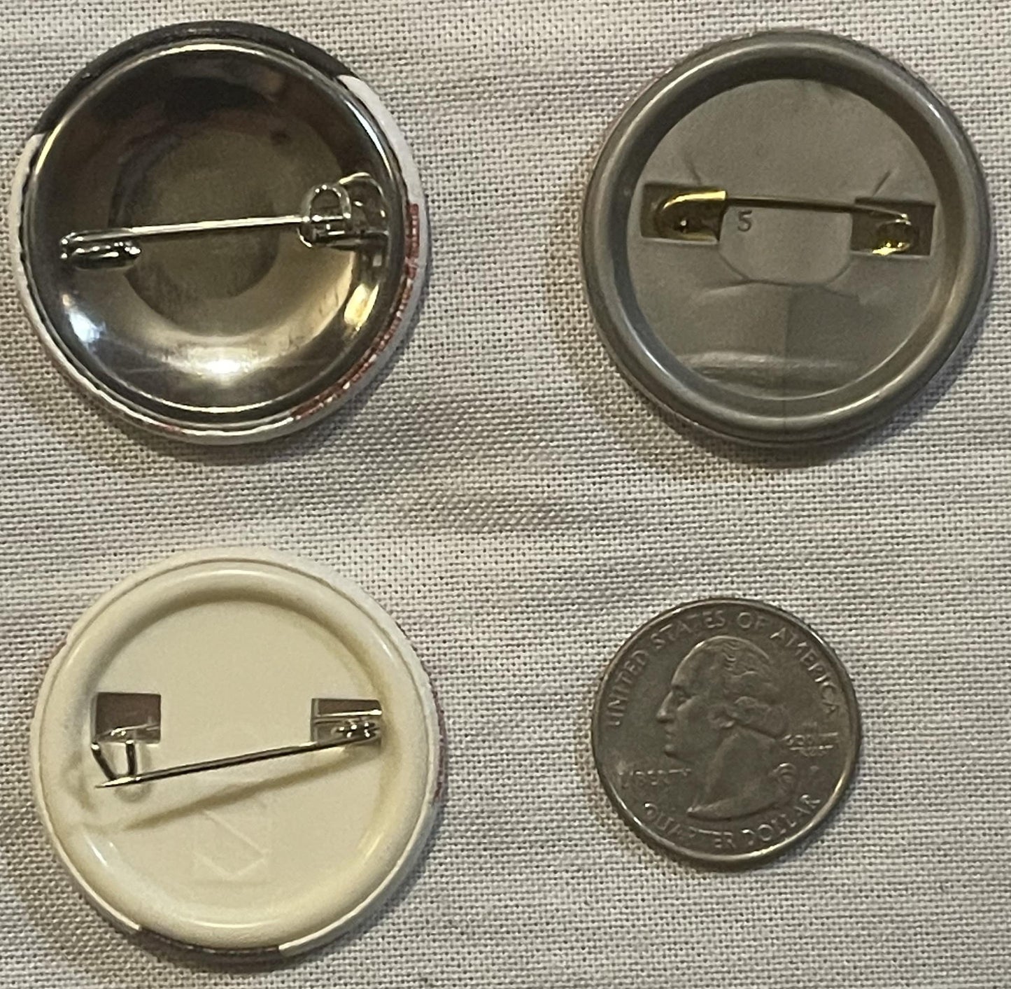 Four pinback buttons displayed with a quarter for scale: two metal versions front and back, one cream-colored version front a