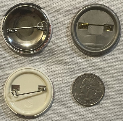 Four pinback buttons shown front and back with a quarter for scale; grotesque crater-faced designs in dark and light finishes
