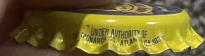 Vintage yellow bottle cap with embossed text reading "Under Authority of Monarch Atman" held by a hand for scale, close-up