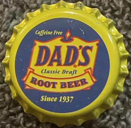 Vintage yellow Dad's Classic Draft Root Beer bottle cap on textured gray fabric, showing blue and red label