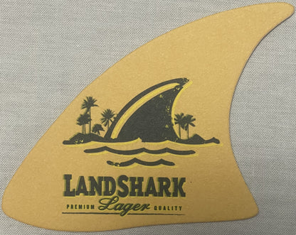 Vintage LandShark Lager shark fin-shaped coaster with gold background featuring black silhouette illustration of shark fin, palm trees, water waves, and "LANDSHARK PREMIUM LAGER QUALITY" text.